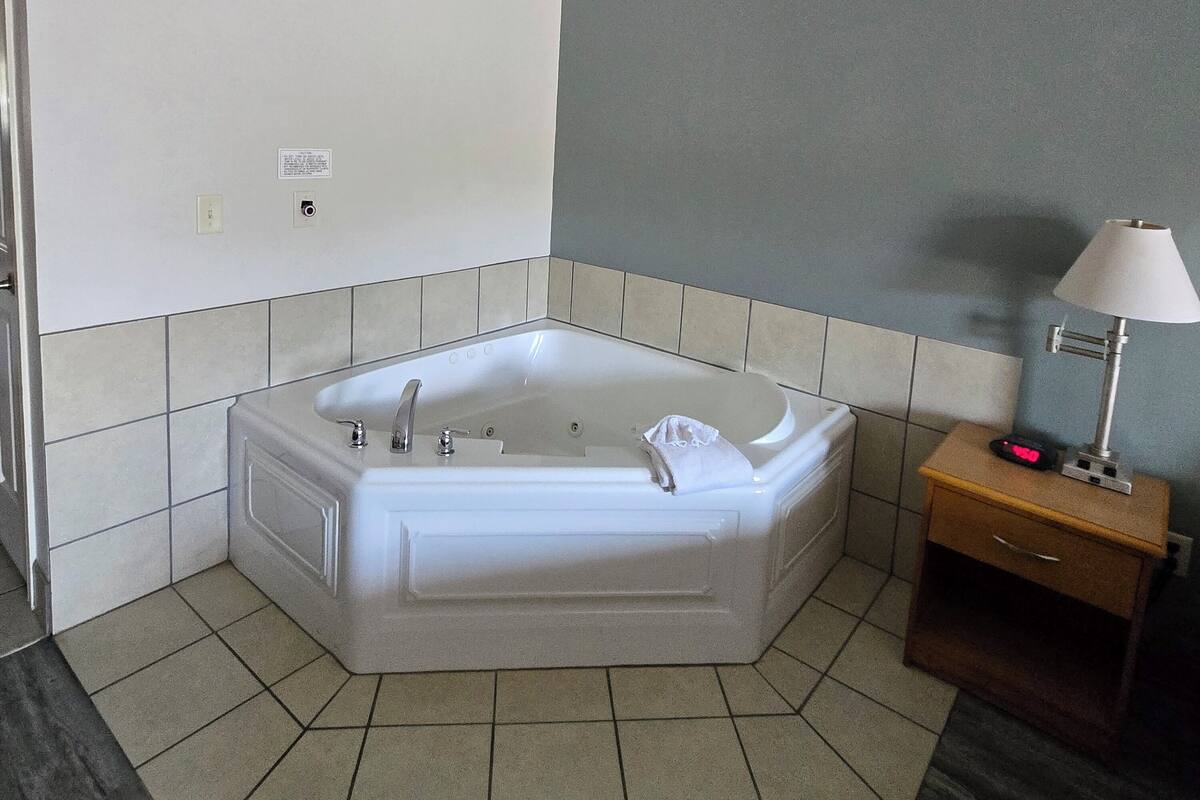 A modern bathroom with a freestanding bathtub, a small bedside table with a lamp, and a tiled floor.