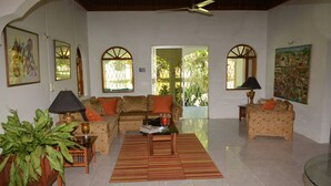 A living room with a sofa, armchairs, a coffee table, and a TV. There are two windows with arched tops, a ceiling fan, and a door leading outside. - Cozy Coco (Negril)
