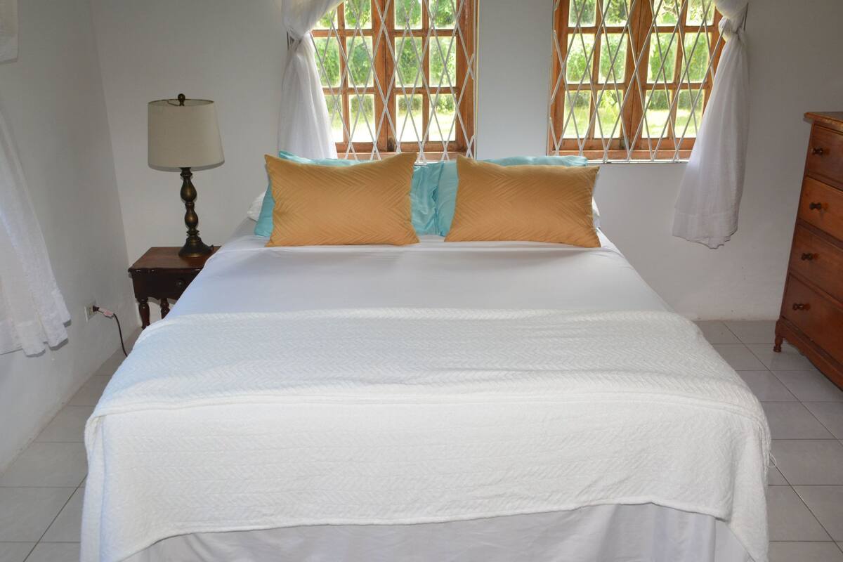 A bedroom with a bed, a nightstand with a lamp, a dresser, and a window with curtains.