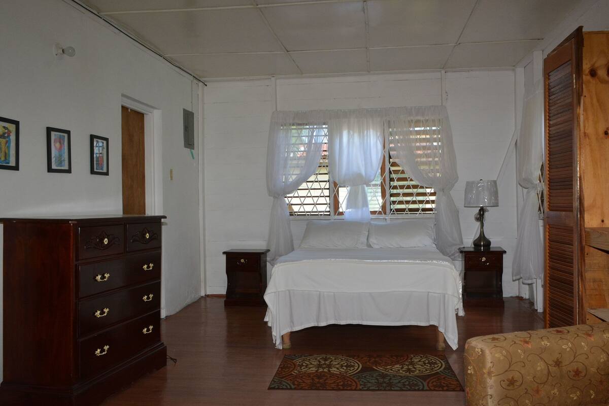 A bedroom with a bed, a wooden dresser, a nightstand, a lamp, and a window with curtains.