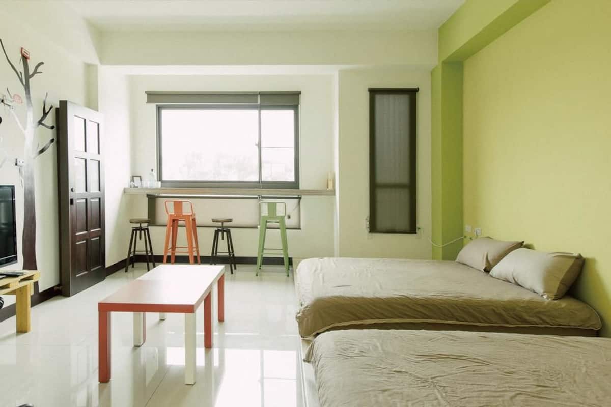 A small, well-lit room with a bed, a desk, and a chair. There is a window providing natural light, and a door is visible in the background.