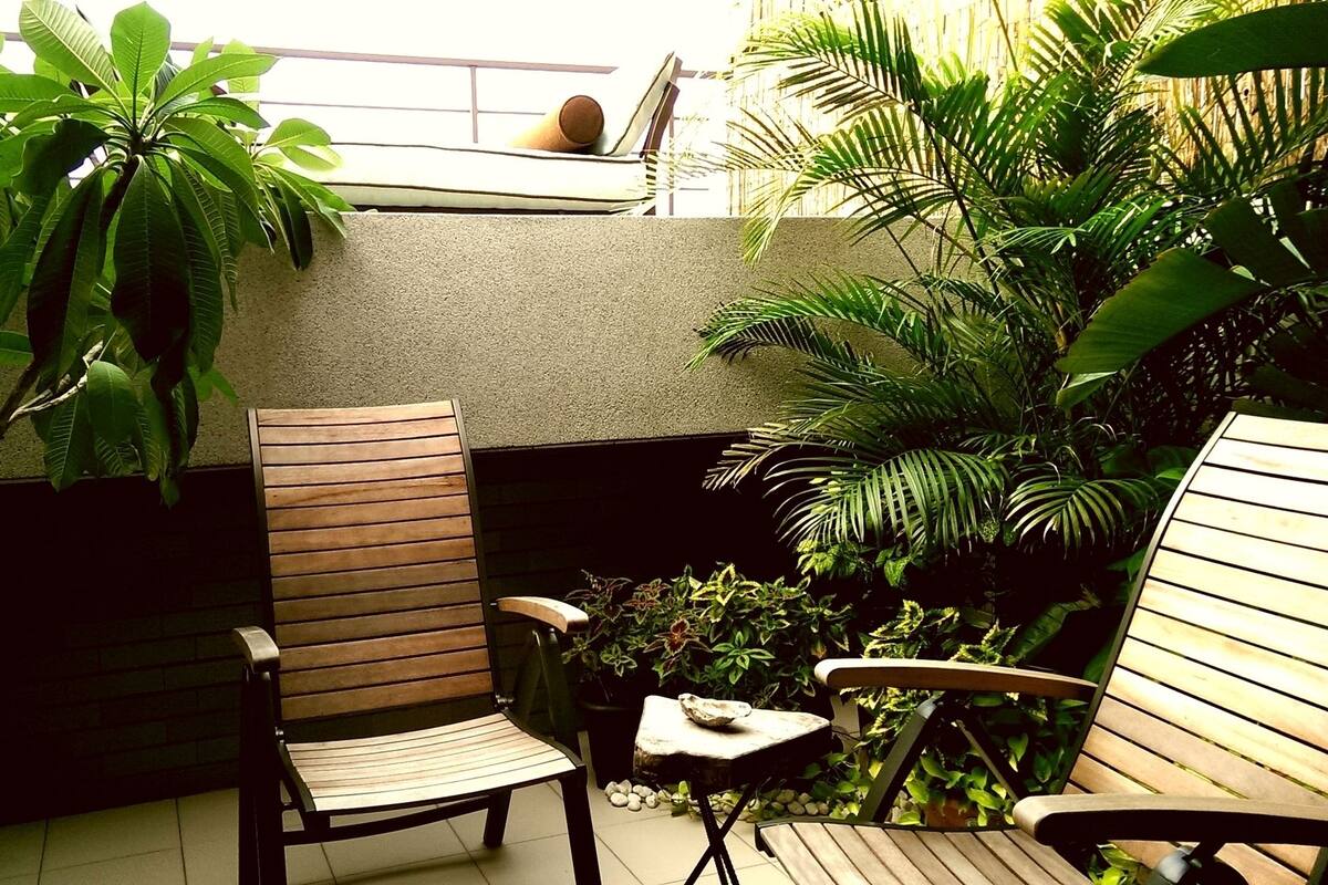 A patio area with wooden chairs and a small table, surrounded by lush green plants and a concrete wall.