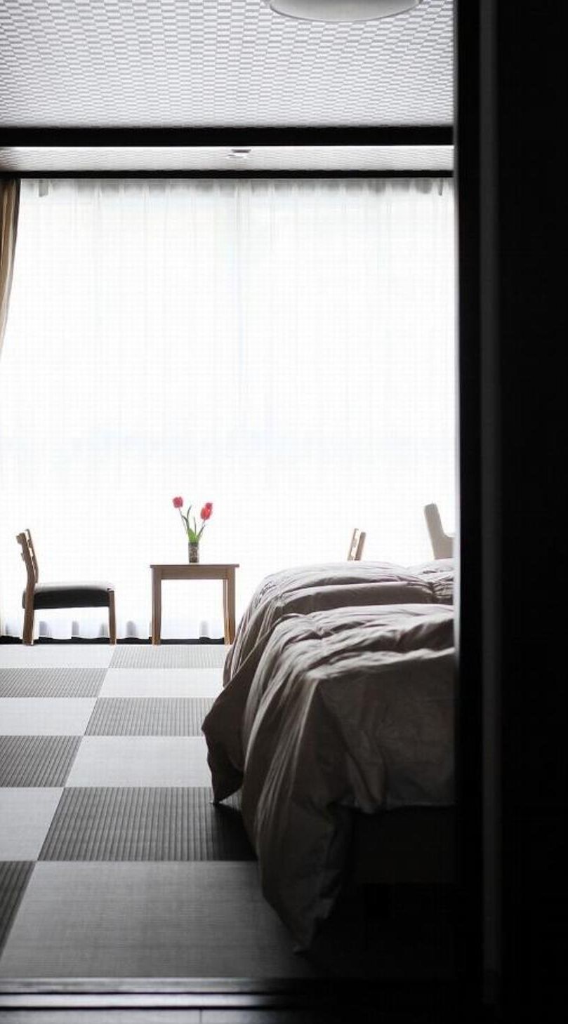 A room with a bed, a chair, a small table with a vase of flowers, and a view through a window.