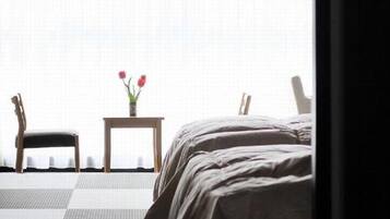 Une pièce comprenant un lit, une chaise, une petite table avec un vase de fleurs et une vue par la fenêtre.