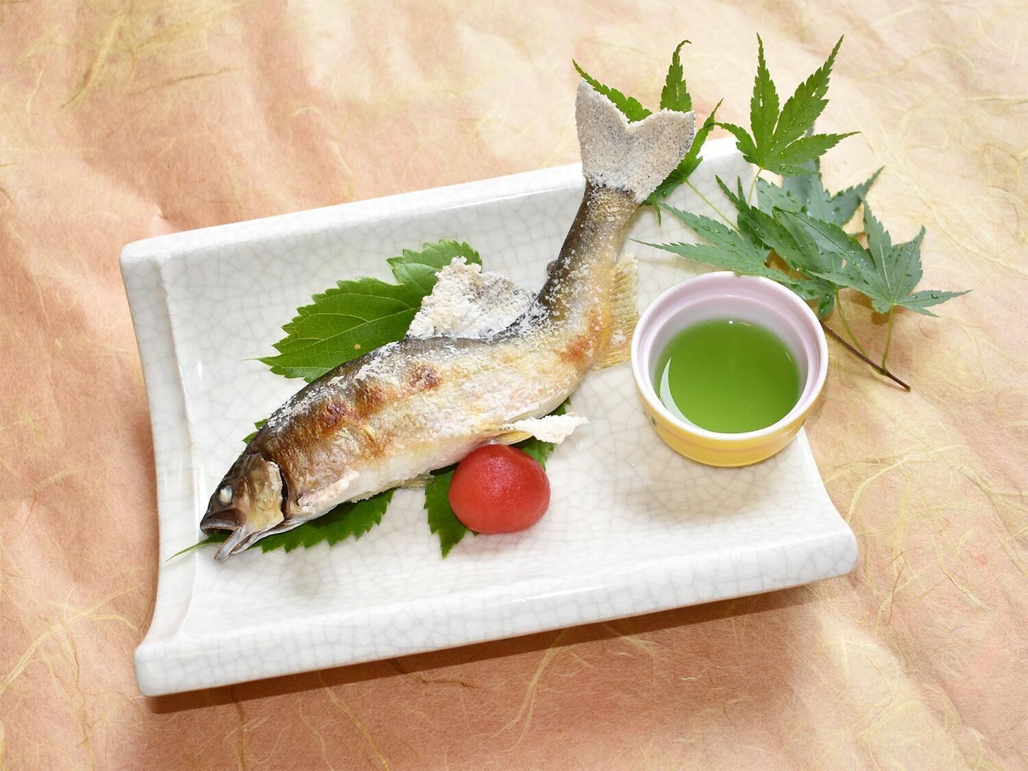 A white rectangular plate with a cooked fish, green leaves, a cherry tomato, and a small bowl of green sauce.