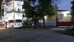 Um edifício de vários andares com portão fechado, uma carrinha branca estacionada em frente e uma pessoa sentada numa mota perto do portão.