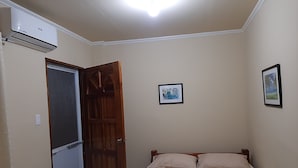 Quarto com cama, porta de madeira, ar-condicionado fixado na parede e duas fotos emolduradas.