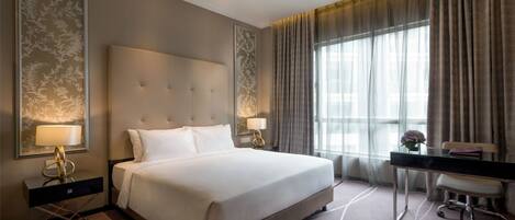 Habitación de hotel con una cama grande, dos mesitas de noche con lámparas, un escritorio con silla y un ventanal con cortinas.