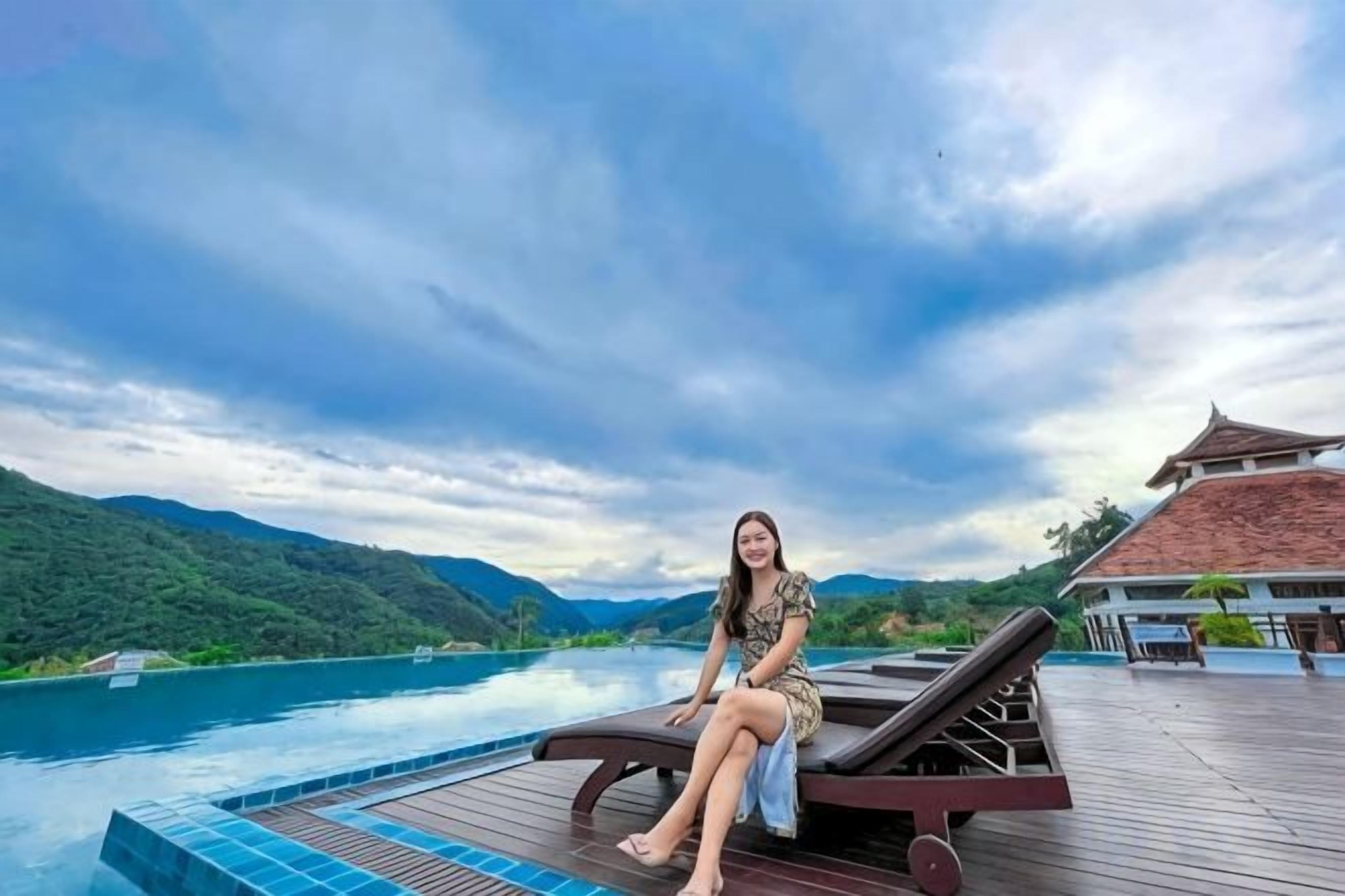 A person sitting by a pool with a wooden deck and a view of a mountain range.