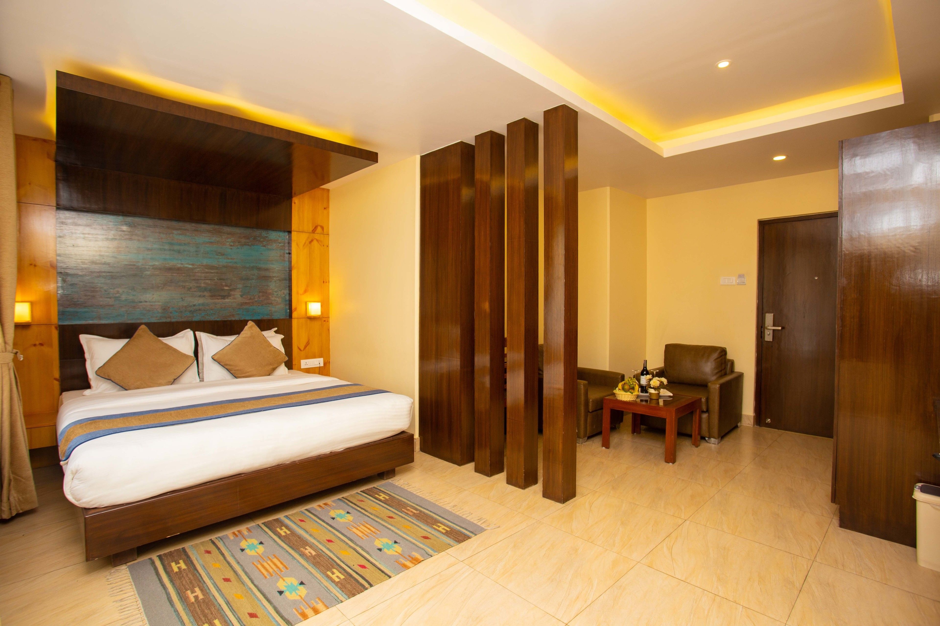 A modern hotel room with a large bed, wooden paneling, a small seating area, and a door.