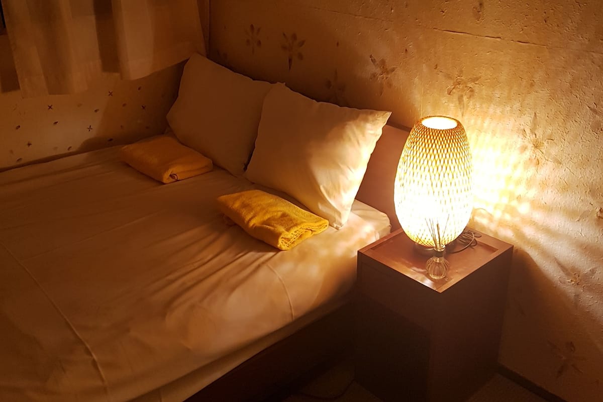 Un lit avec du linge de lit blanc, deux oreillers et une serviette jaune. Il y a une table de chevet avec une lampe à côté du lit.