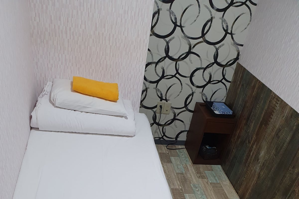 Une petite chambre avec un lit simple, un plancher en bois, un mur décoré et une petite table de chevet en bois sur laquelle se trouve un ordinateur portable.