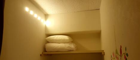 Una habitación pequeña y tenuemente iluminada con una litera, un estante con almohadas y un adorno en la pared.