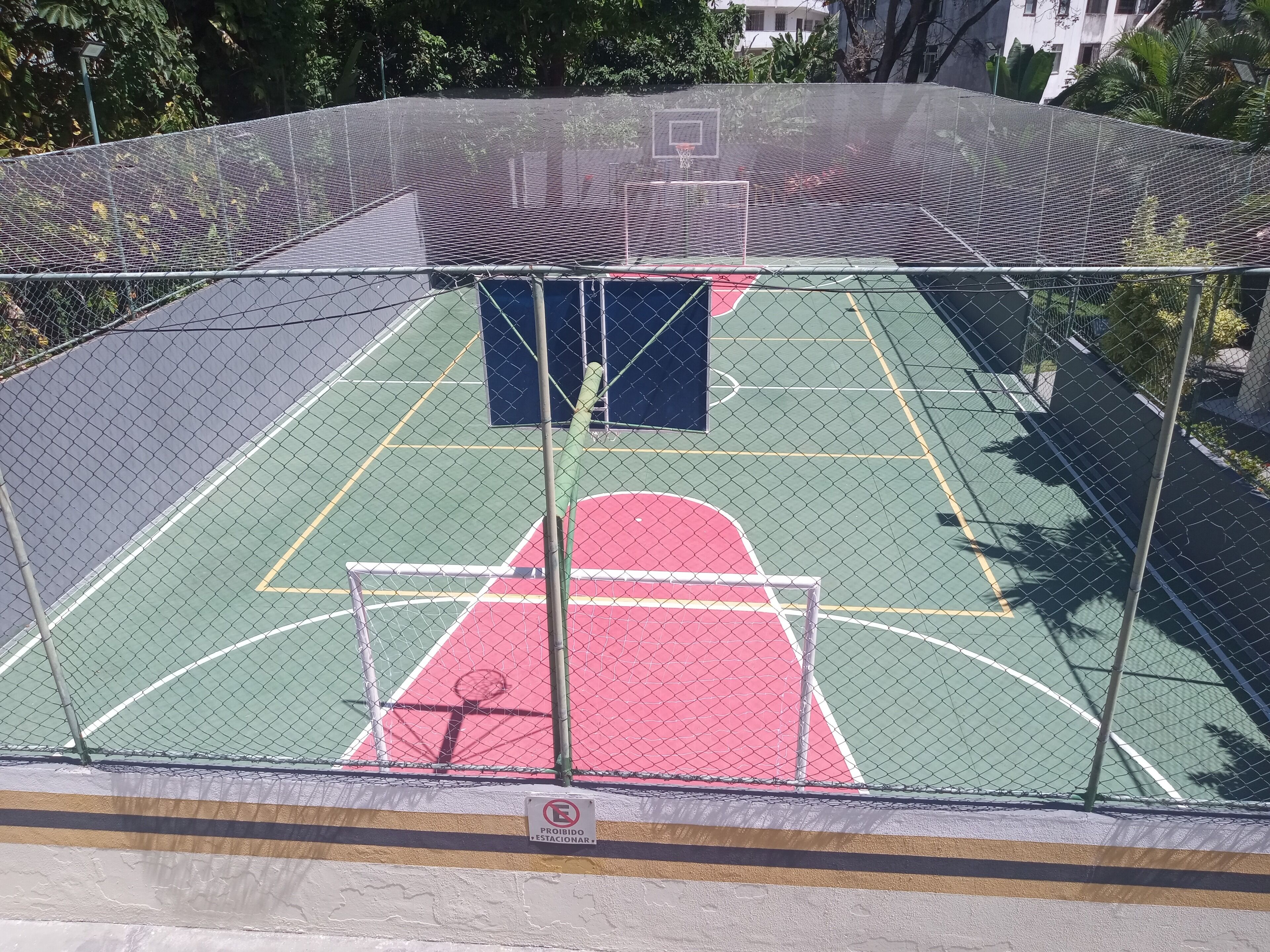 A basketball court with a chain-link fence, a basketball hoop, and a "Prohibido Estacionar" sign.