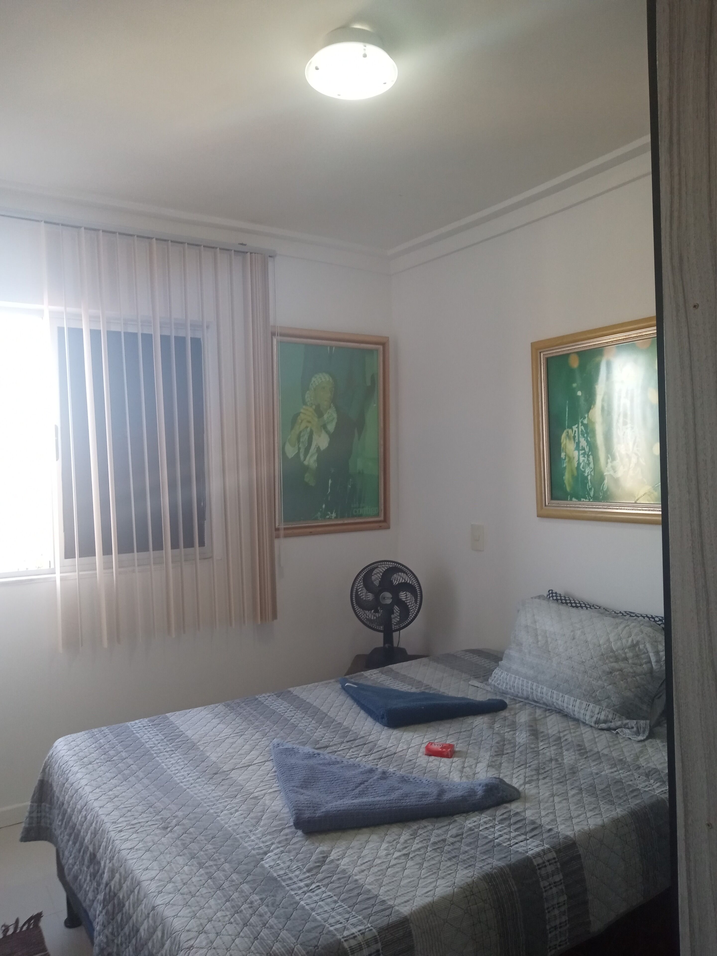 A bedroom with a bed, a standing fan, and two framed pictures on the wall.