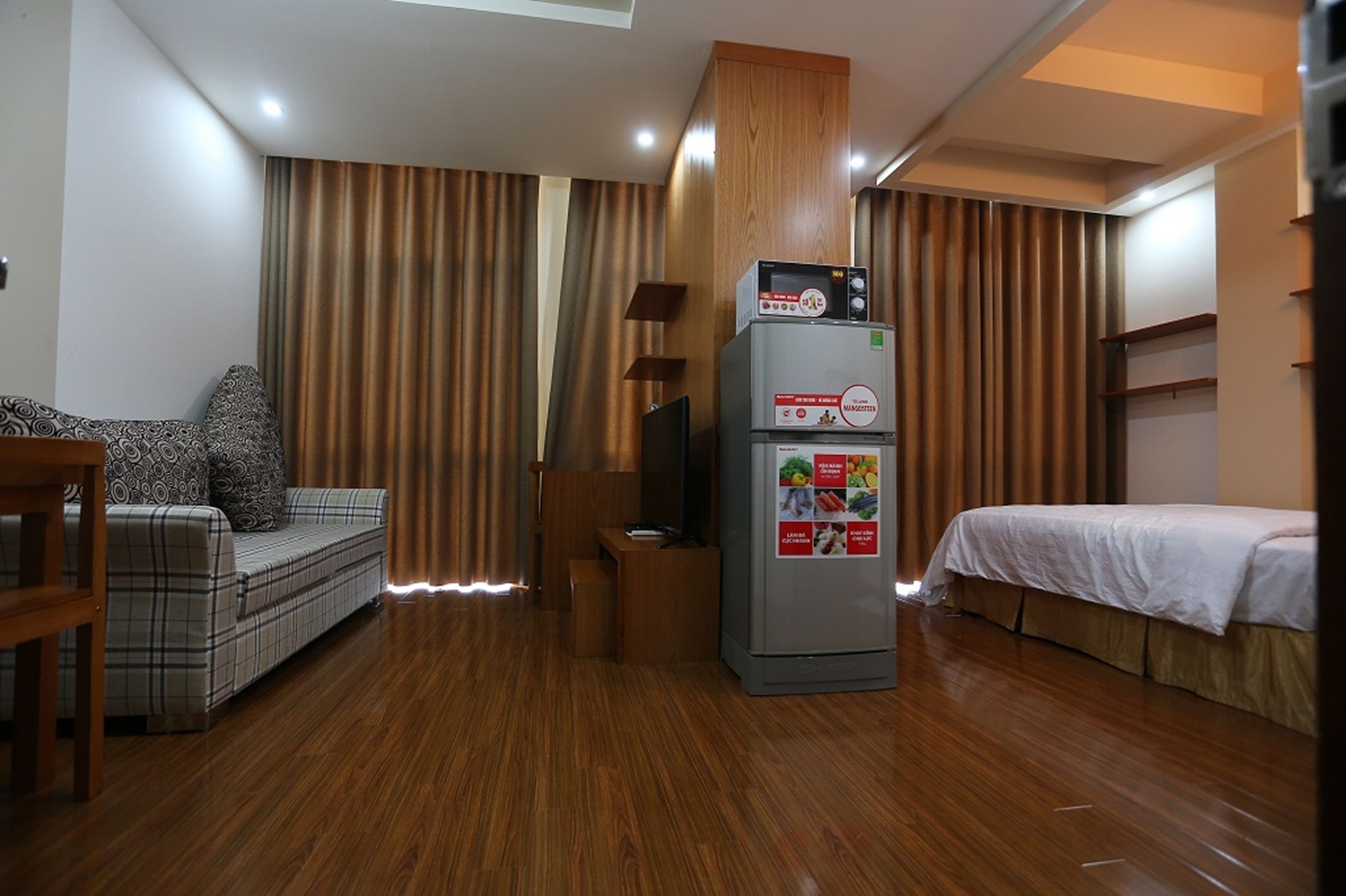 A compact hotel room with a sofa, a bed, a refrigerator, and wooden flooring.