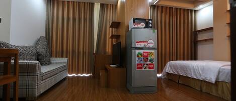 A compact hotel room with a sofa, a bed, a refrigerator, and wooden flooring.