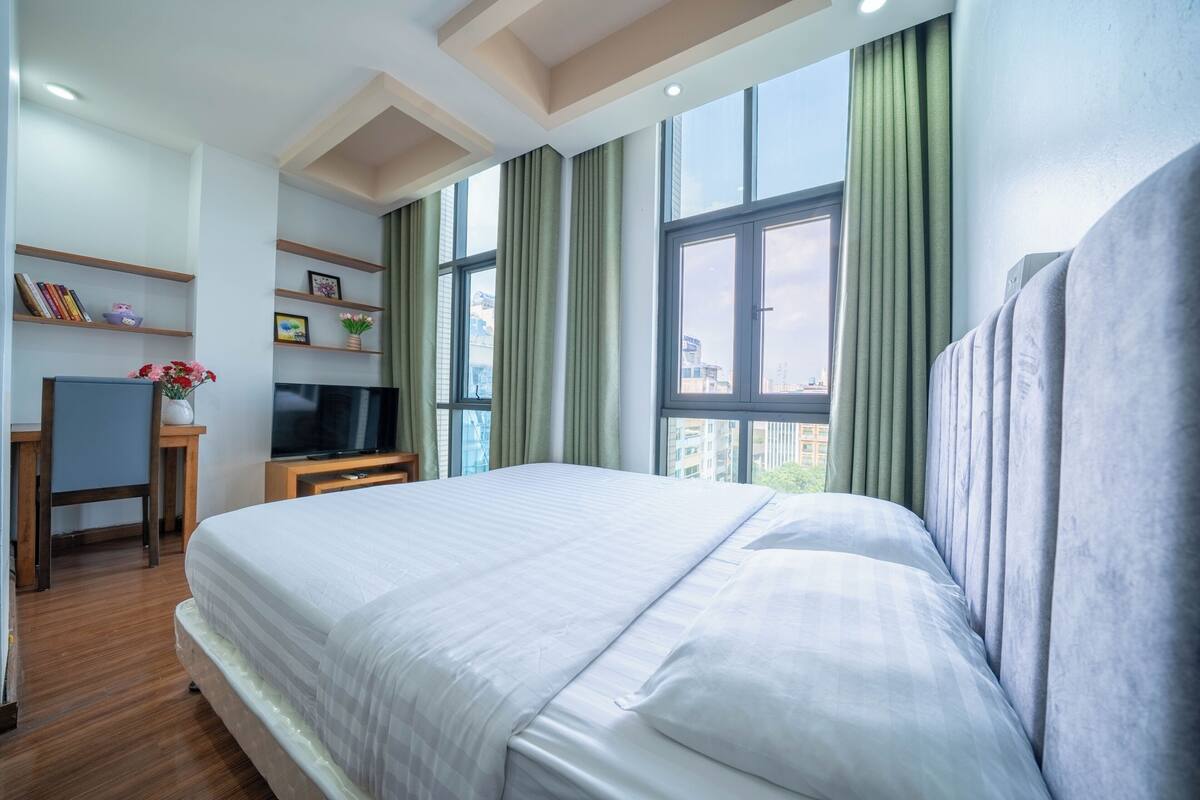 A hotel room with a large bed, a desk with a TV, shelves with books and flowers, and large windows with curtains.