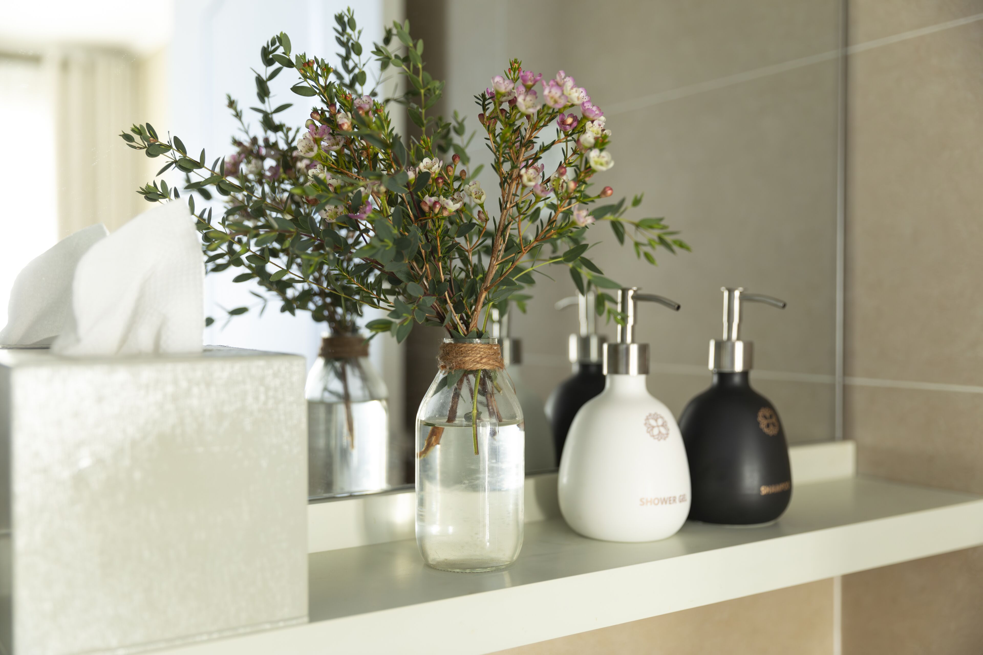 A bathroom shelf with a clear glass vase containing flowers, a white soap dispenser, and a black soap dispenser.