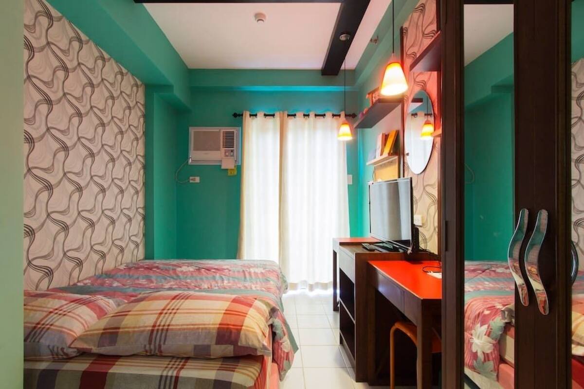 A room with teal walls, two beds with plaid bedding, a desk with a computer, and a mirror.