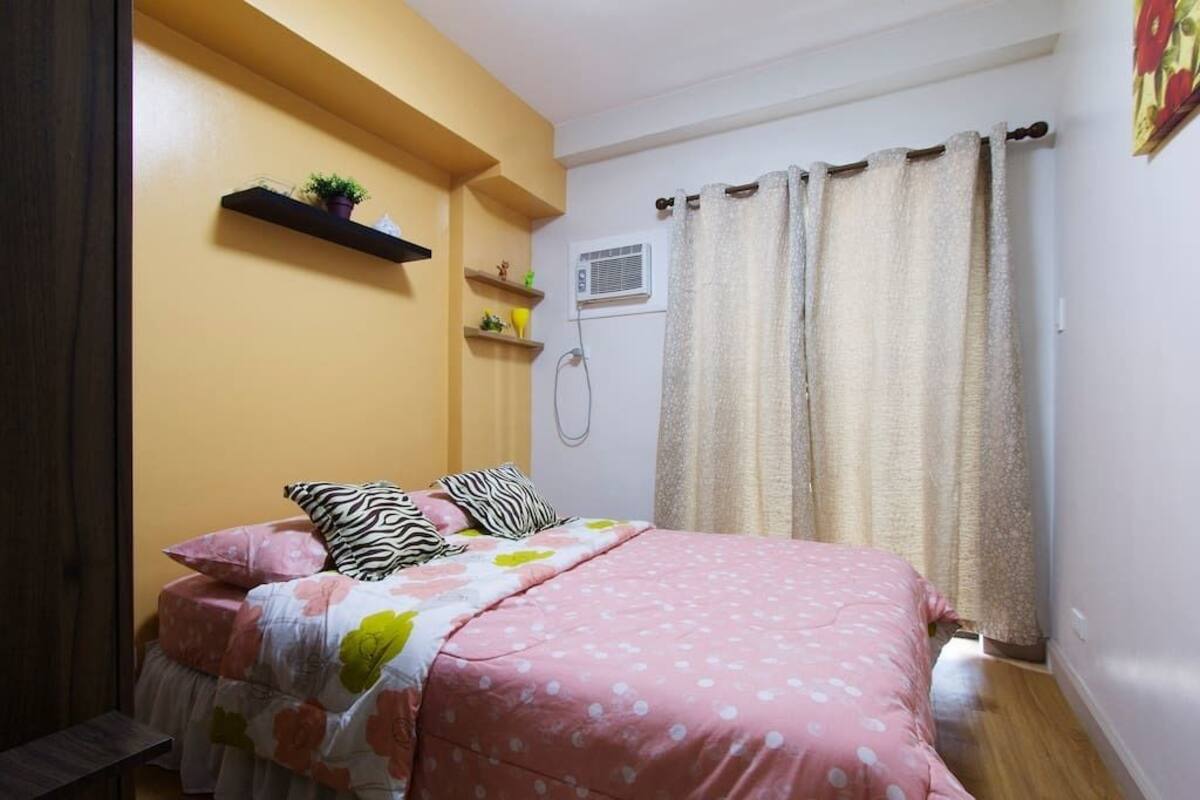 A bedroom with a bed, a shelf with a plant, an air conditioning unit, and a shower curtain.