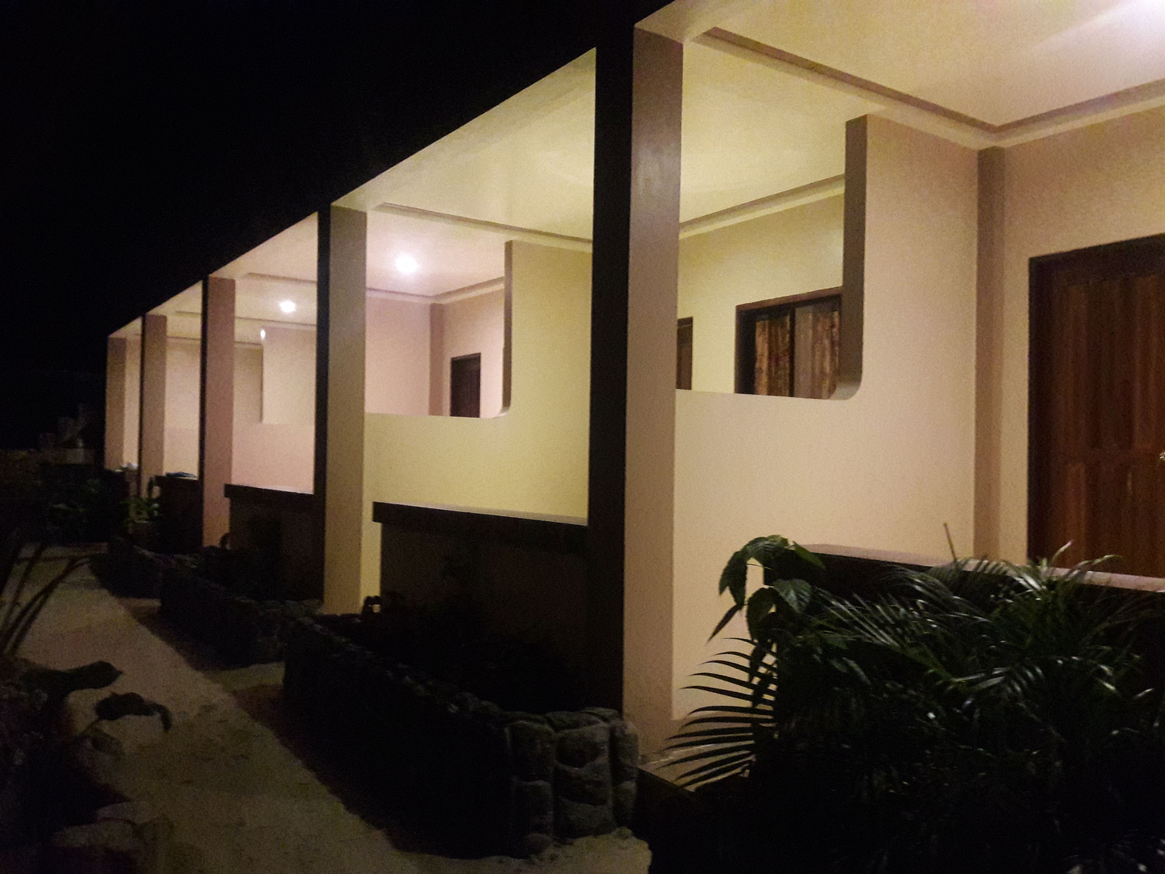 Un edificio con varias habitaciones, cada una con una puerta y una ventana pequeña visibles. Hay plantas en macetas afuera, y el edificio tiene techo plano.