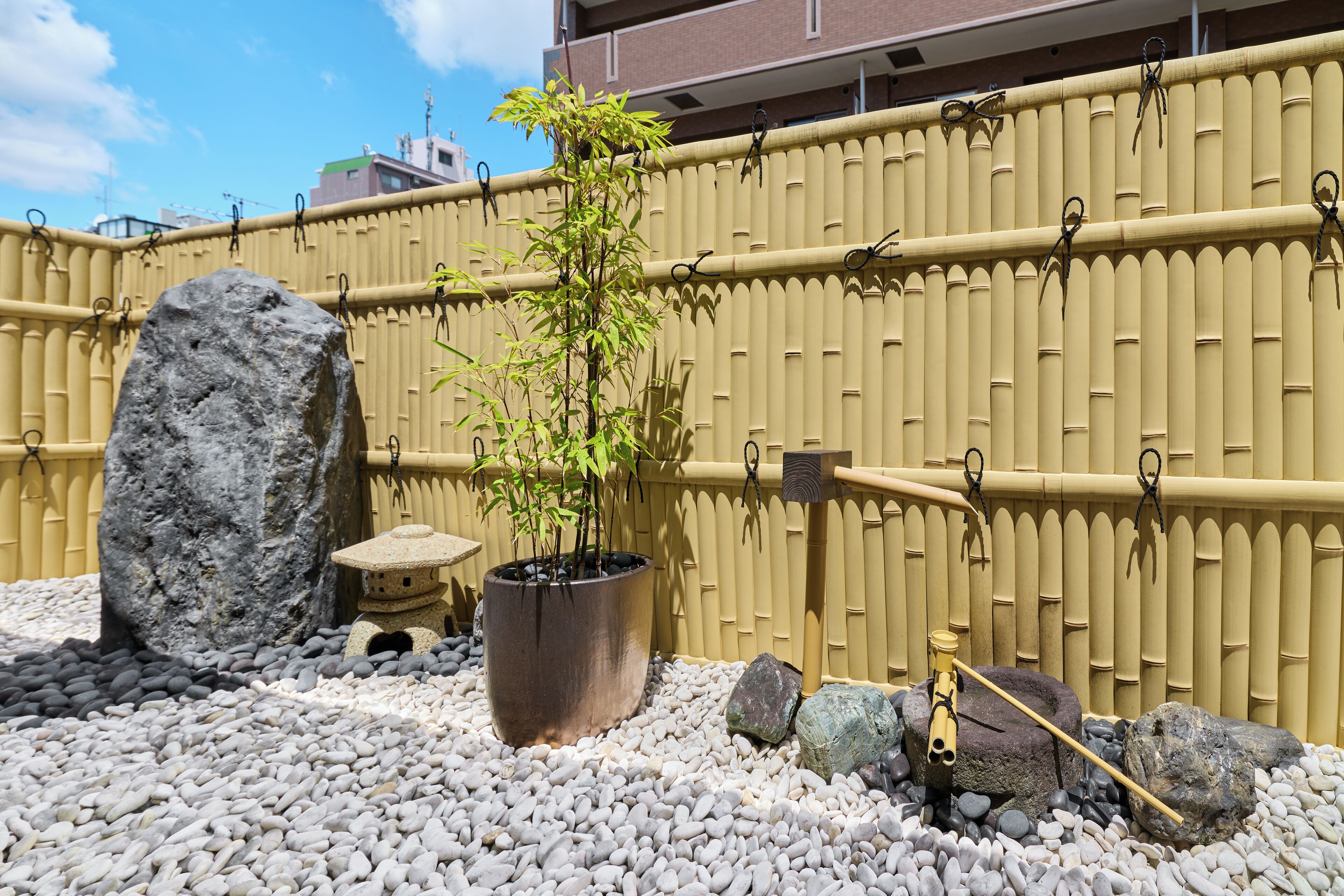 A garden with a bamboo fence, a potted plant, rocks, and a traditional Japanese water feature.