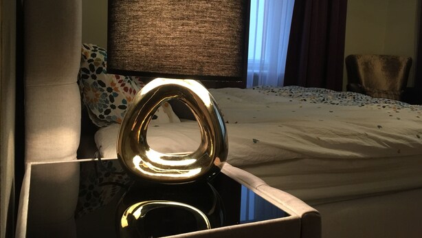 A bed with a white comforter, a round metallic object on a glass table, a lamp with a textured shade, and a chair in the background. - Heart of Riga Apartment (Riga)