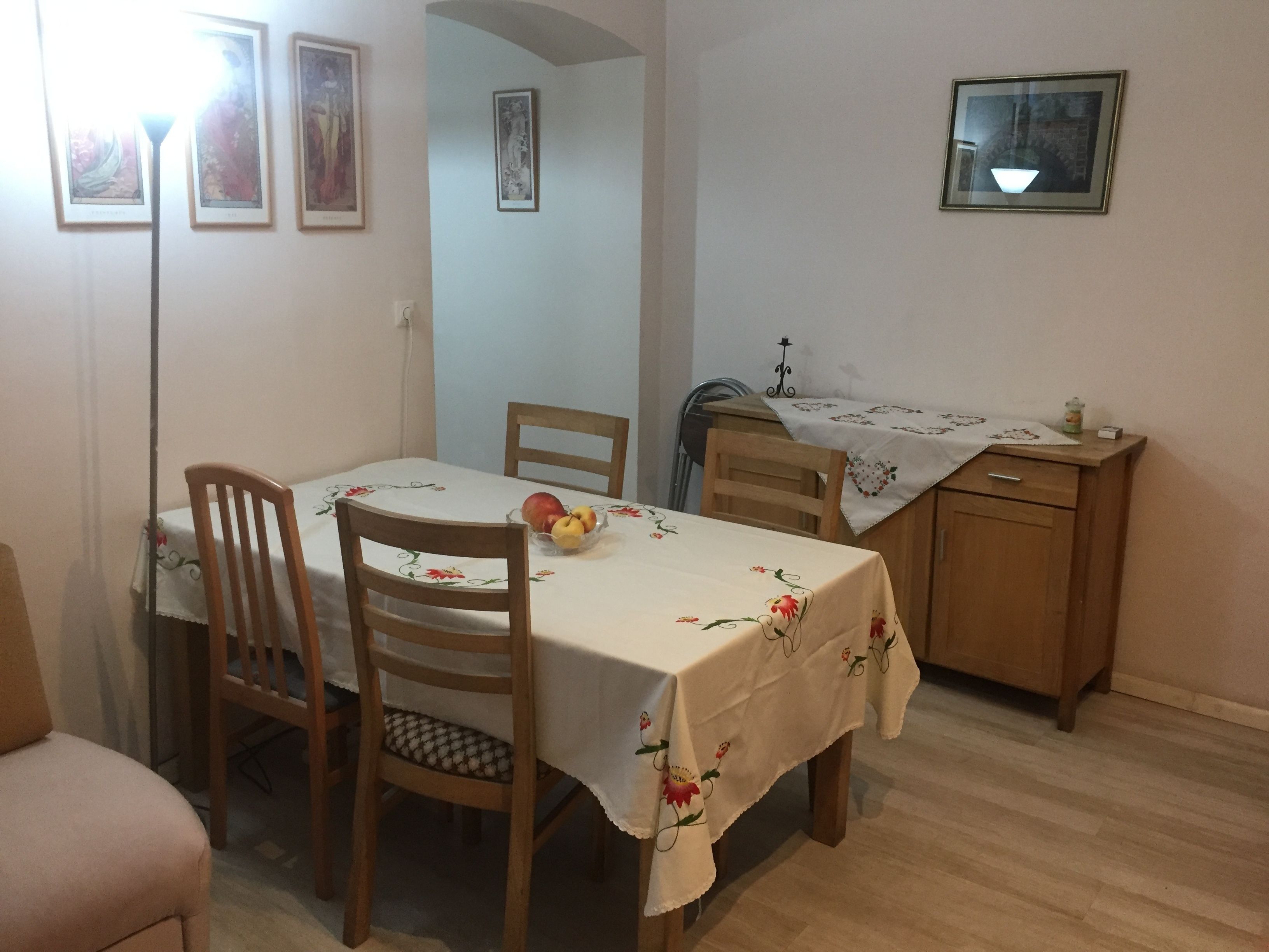 Un área de comedor con una mesa dispuesta para dos personas, sillas de madera, un frutero pequeño y un aparador con un paño decorativo.
