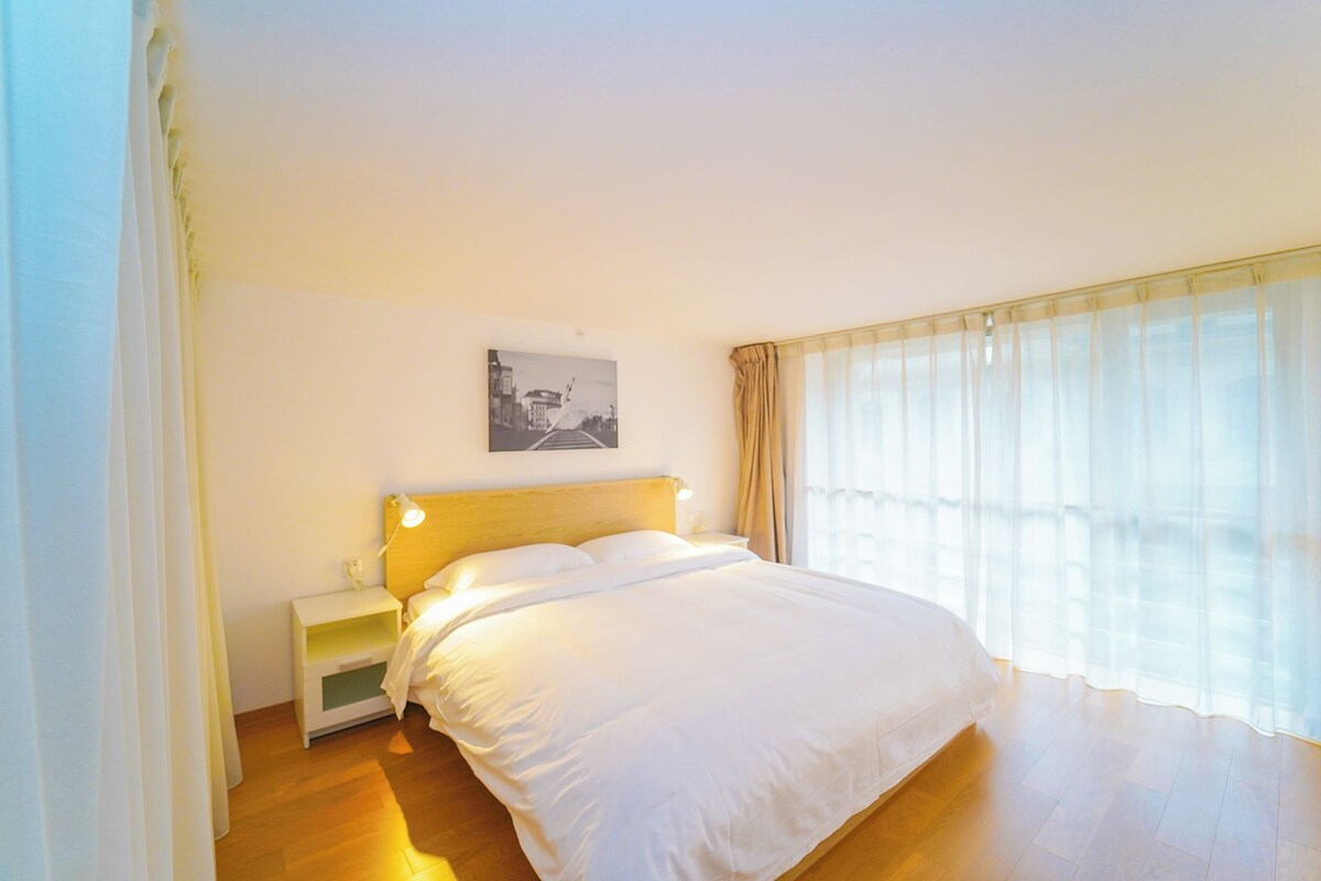 A hotel room with a large bed, a nightstand, a wall-mounted lamp, a window with sheer curtains, and a framed picture on the wall.