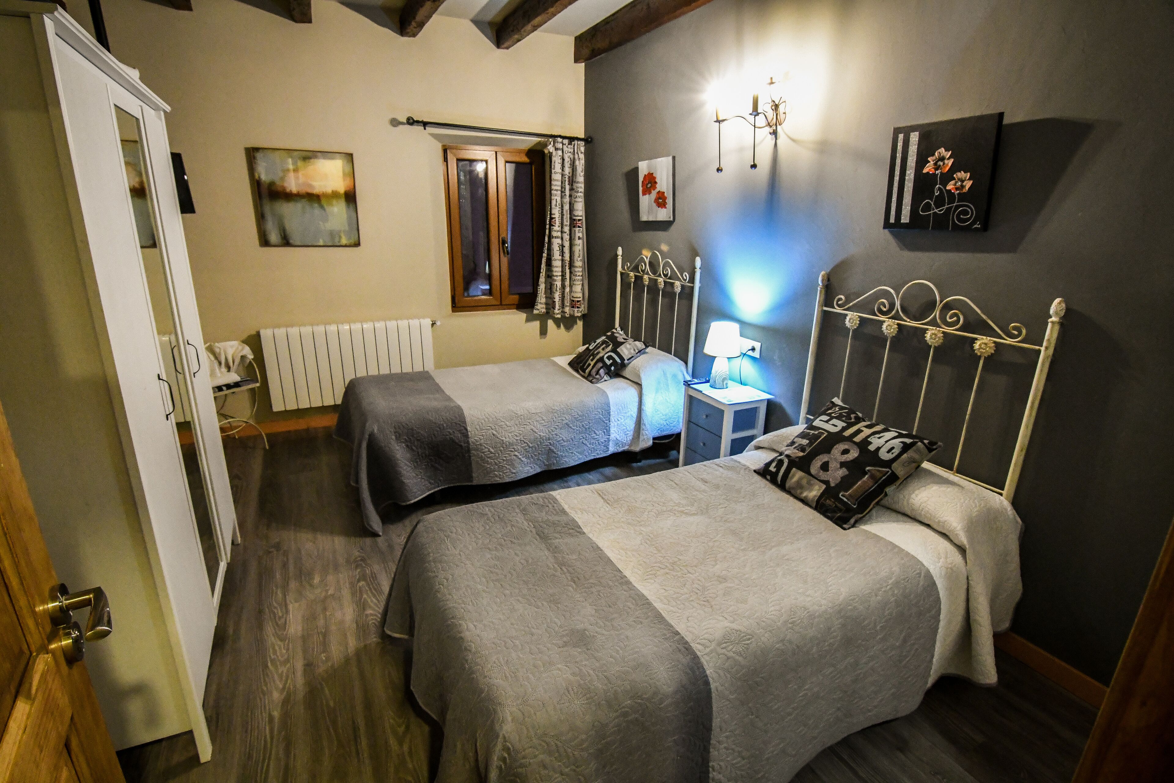 A bedroom with two beds, a nightstand, a lamp, a window with curtains, and a radiator.