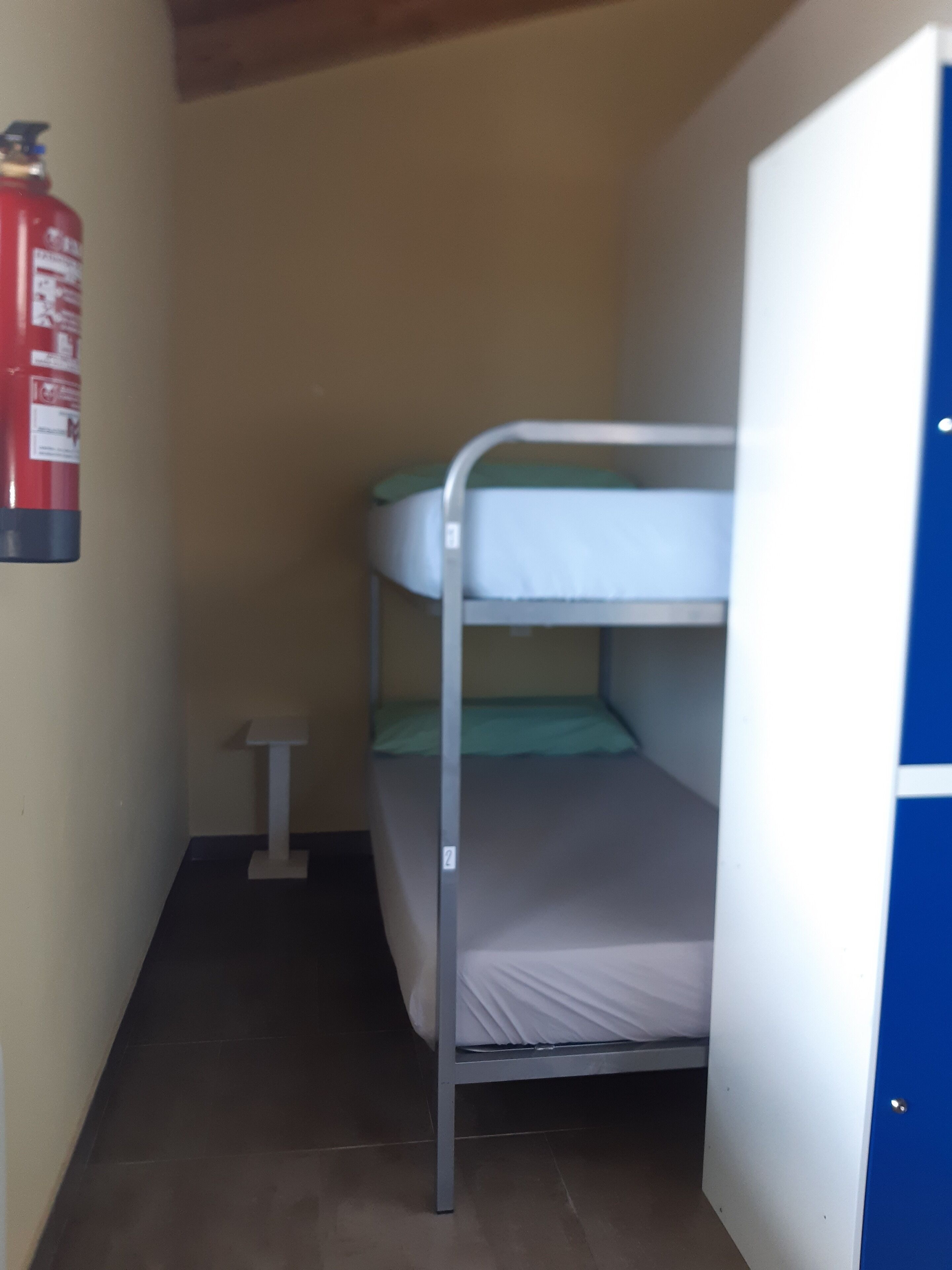 Ett litet, smalt rum med en våningssäng, ett vitt bord och en brandsläckare på väggen.