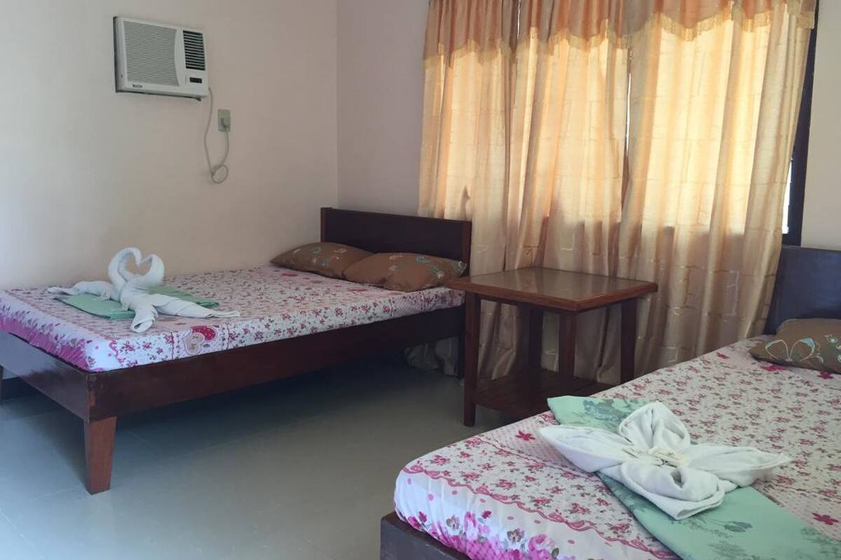A hotel room with two single beds, a small table, a chair, a wall-mounted air conditioner, and a window with curtains.