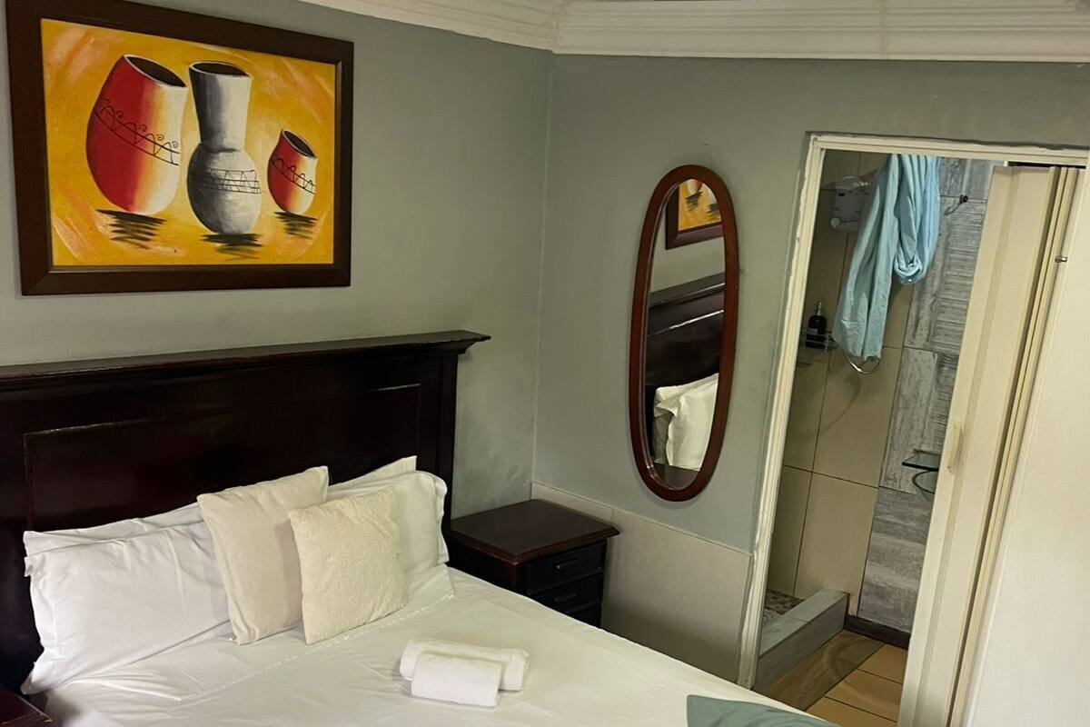 Un lit bien fait, avec du linge de lit blanc et un détail turquoise, une table de chevet en bois, une petite table, un tableau représentant des vases et un miroir au mur.