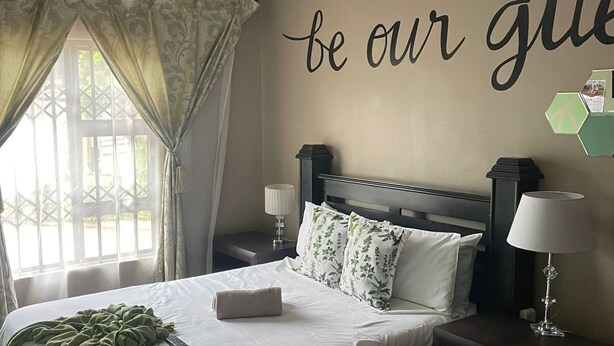 Un lit bien fait, avec une couverture verte et une télécommande, une table de chevet avec une lampe, et un mur orné d’une citation inspirante.