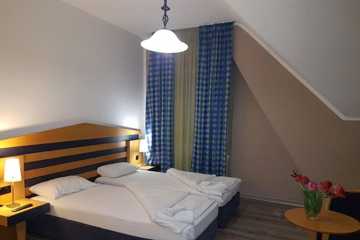 A hotel room with a double bed, a nightstand with a lamp, a small table with a vase of flowers, and a view of the outside through a window with blue curtains.
