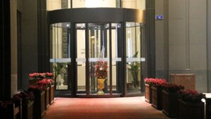 Une porte tournante marque l’entrée, précédée d’un tapis rouge et bordée de plantes en pot ornées de fleurs rouges.