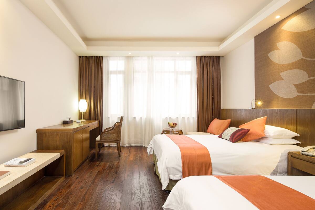 Habitación de hotel con un escritorio de madera, dos camas, un televisor y un amplio ventanal con cortinas.