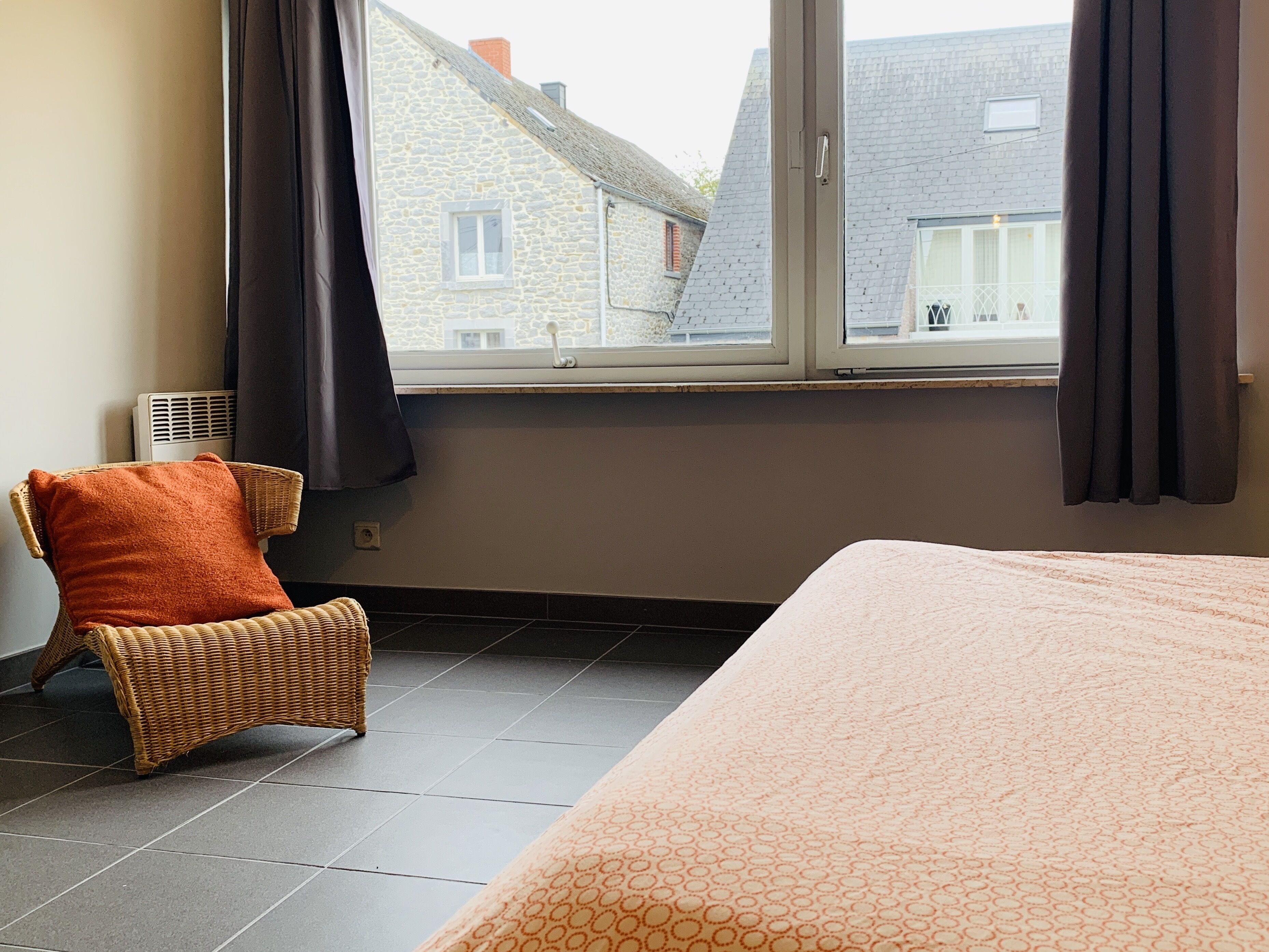 A bedroom with a bed, a wicker chair, a window with curtains, and a view of buildings outside.