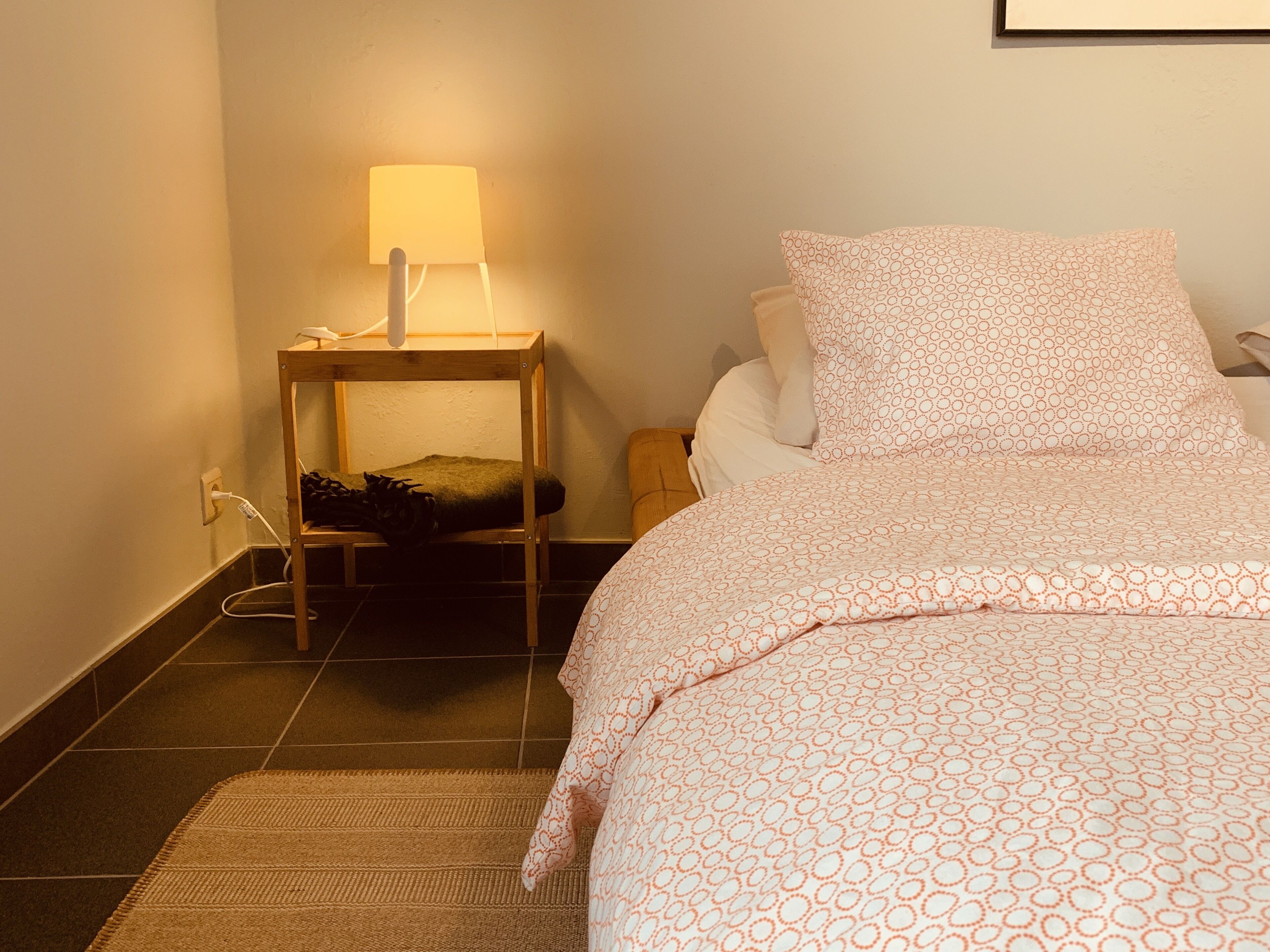 Een net opgemaakt bed met een beddekleed met patroon, een bedlampje en een klein houten nachtkastje met een oplader.