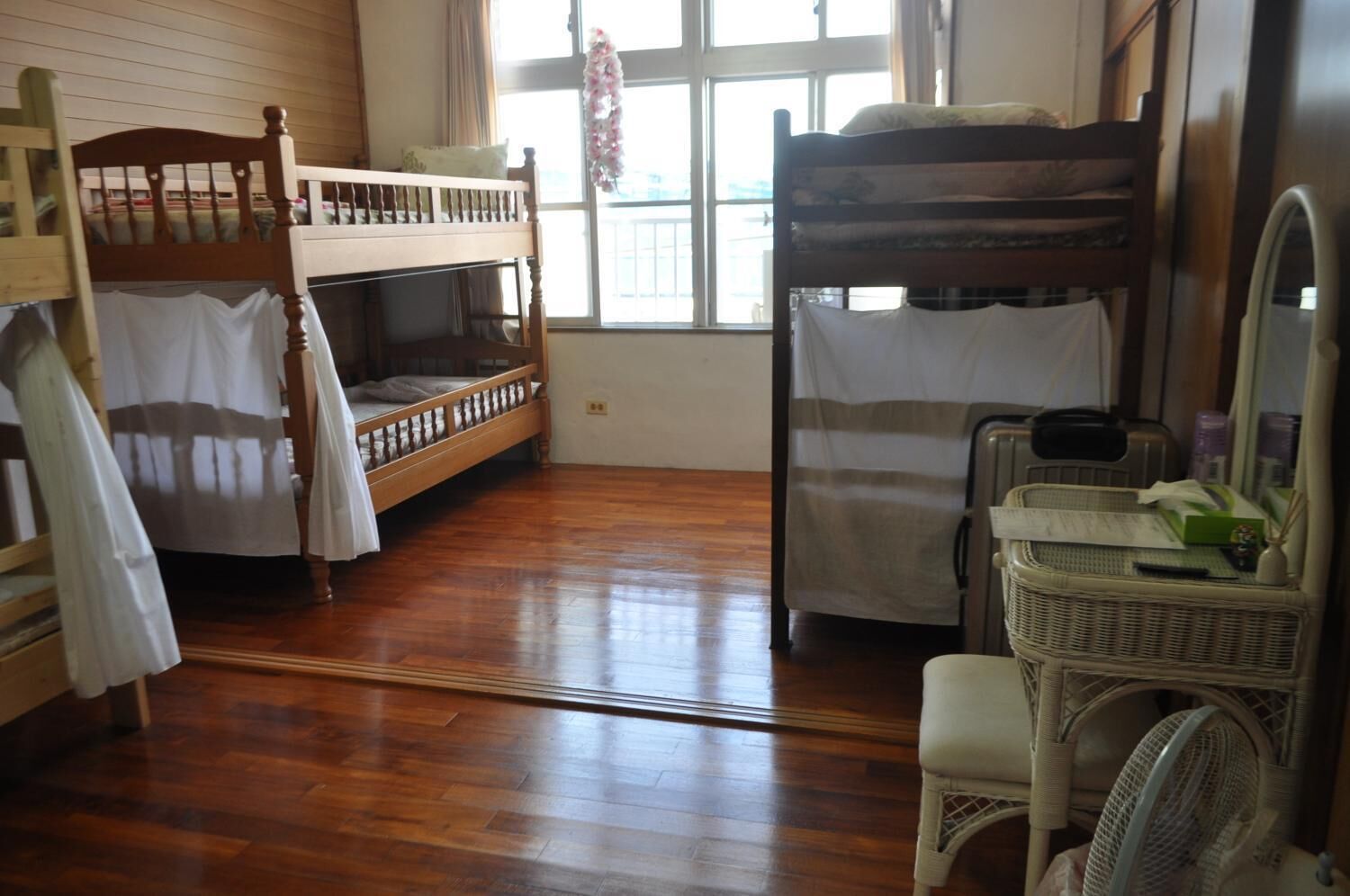 A dormitory room with two bunk beds, a wicker chair, a desk with a fan, and a window with curtains.
