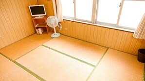 Una habitación con esteras de tatami, un televisor, un ventilador y una ventana con cortinas.