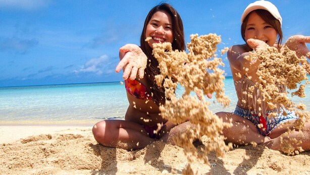 Dos personas sentadas en una playa arenosa con un cielo azul despejado y el océano al fondo.