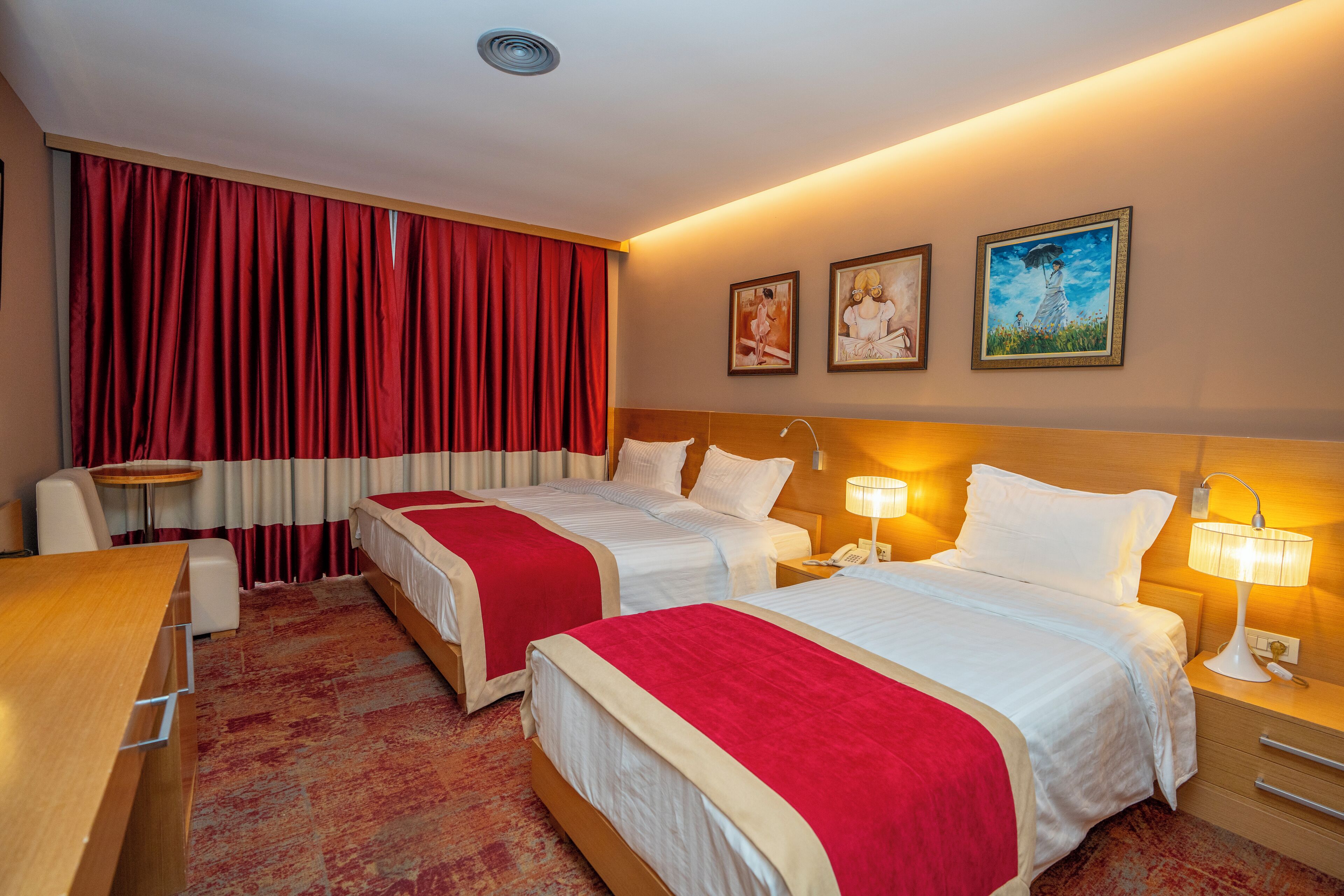 A hotel room with two beds, a desk, a chair, and a small table. There are three framed pictures on the wall.