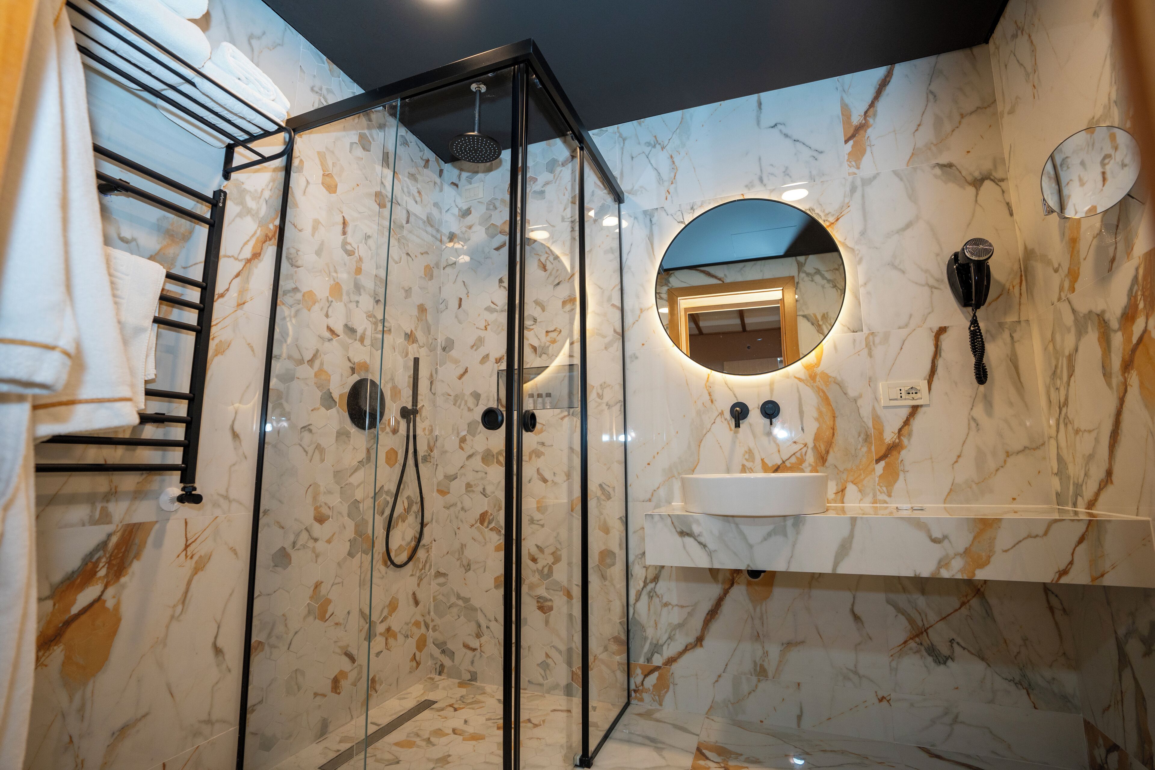A modern bathroom with a glass-enclosed shower, marble walls, a round mirror, and a white sink.