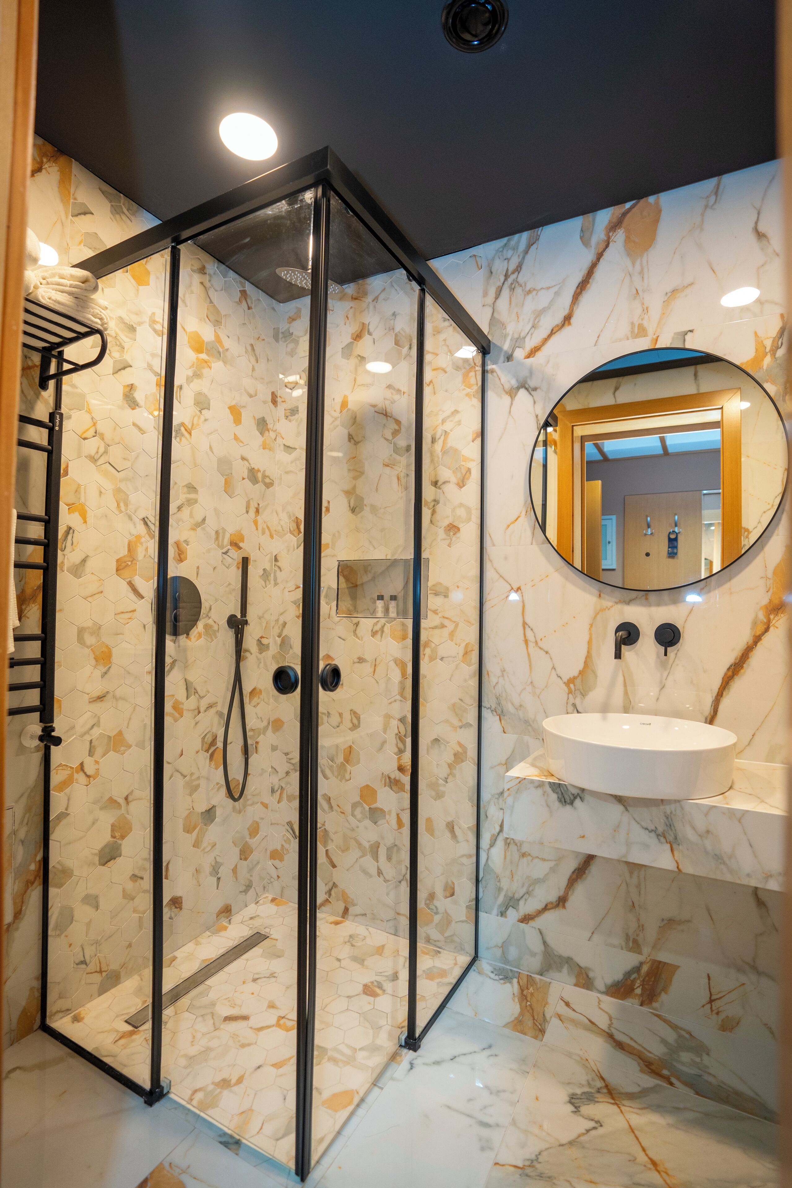 A modern bathroom with a glass-enclosed shower, a marble-tiled wall, a round mirror, and a white sink.