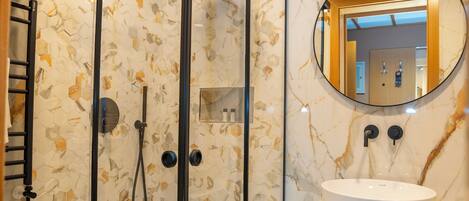 A modern bathroom with a glass-enclosed shower, a marble-tiled wall, a round mirror, and a white sink.