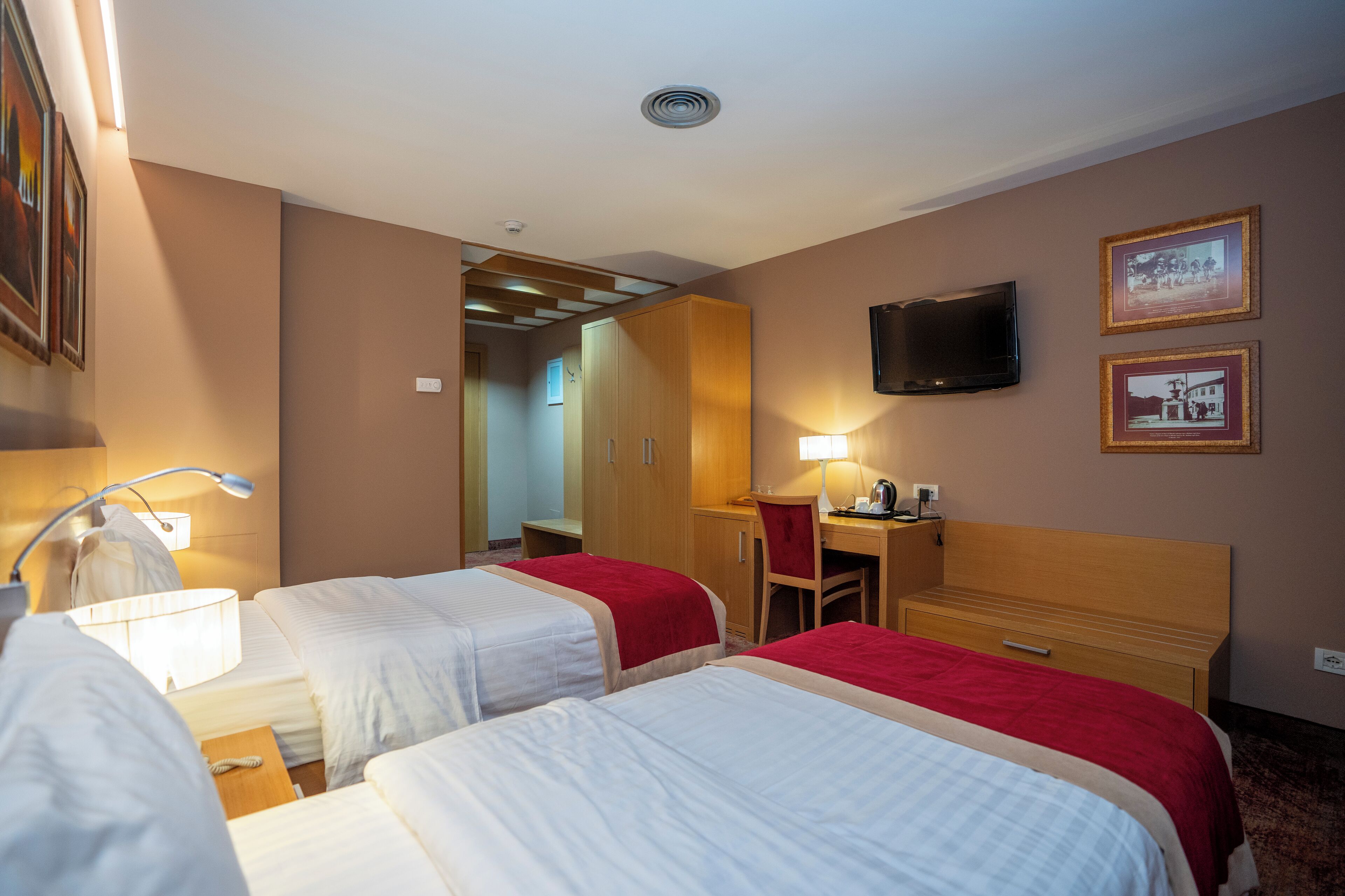 Hotellihuone, jossa on kaksi sänkyä, työpöytä, tuoli, televisio ja seinällä kehystettyjä tauluja.