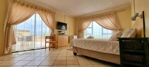 Une chambre d’hôtel équipée d’un lit, d’un bureau, d’une chaise, d’une télévision et de grandes fenêtres avec des rideaux.