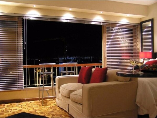 A balcony with a view of a cityscape at night, a sofa with red cushions, and a small table with a lamp.